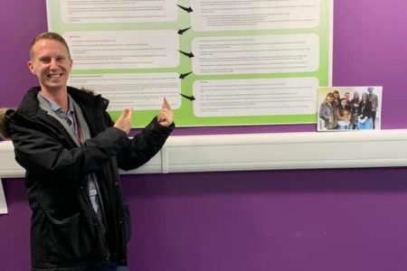 student daniel pointing at his student-staff partnership project poster on the wall