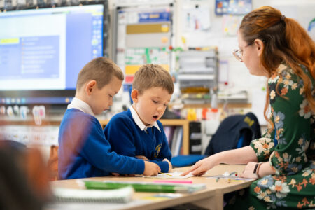 Teacher sat at desk with primary pupils pointing at workbook