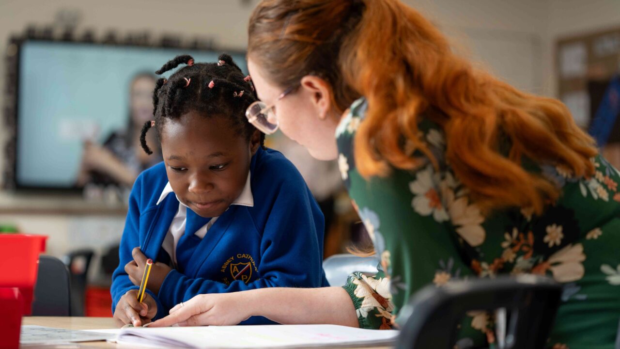 Teacher sat at desk with primary pupil pointing at workbook