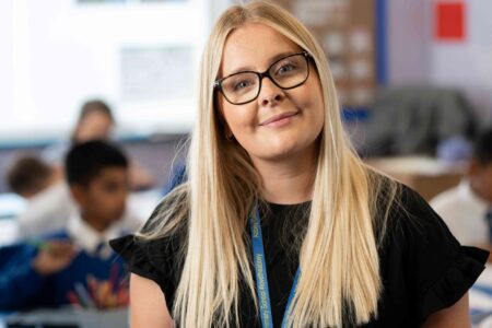 Primary teacher facing camera with pupils in a classroom in the background