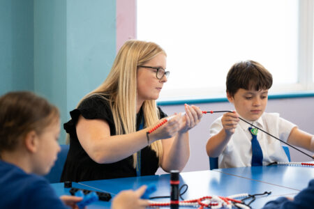 Teacher putting beads on a string assisted by a young pupil